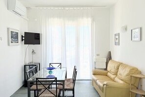 A living room with a glass dining table, a beige sofa, a TV mounted on the wall, and a window with sheer curtains.