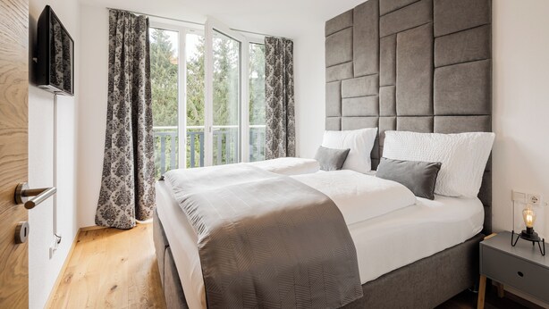 A bedroom with a large bed, a flat-screen TV mounted on the wall, a bedside table with a lamp, and a window with curtains. - Exclusive apartment with mountain views in a ski paradise (Mauterndorf)
