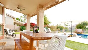 An outdoor patio with a wooden table and white chairs, a ceiling fan, and a view of a swimming pool and garden.