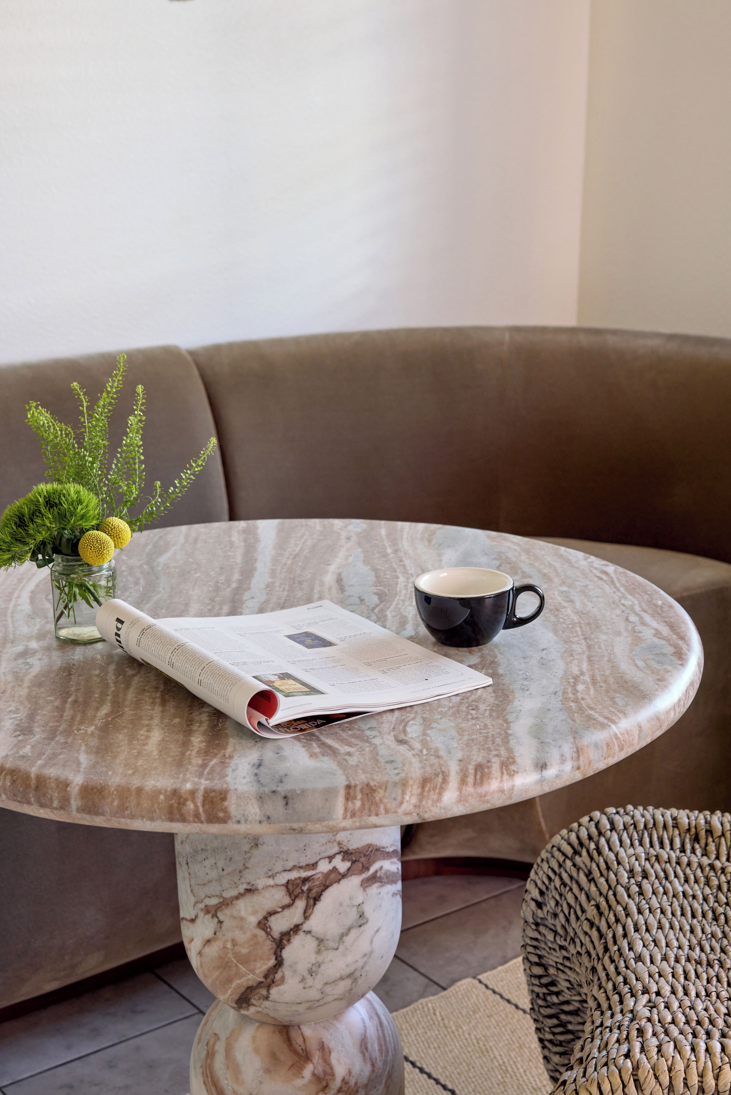 A marble table with a book and a cup, a vase with green plants, and a brown sofa in the background.