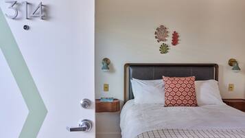 A hotel room with a bed, a nightstand, a wall-mounted light, and a door with a number.