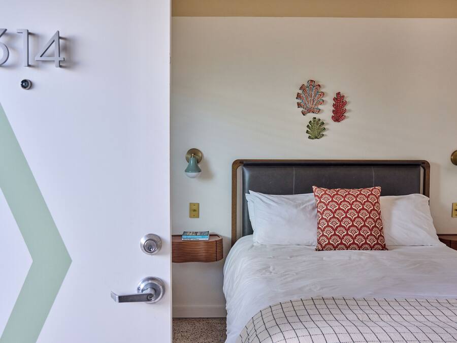 A hotel room with a bed, a nightstand, a wall-mounted light, and a door with a number.