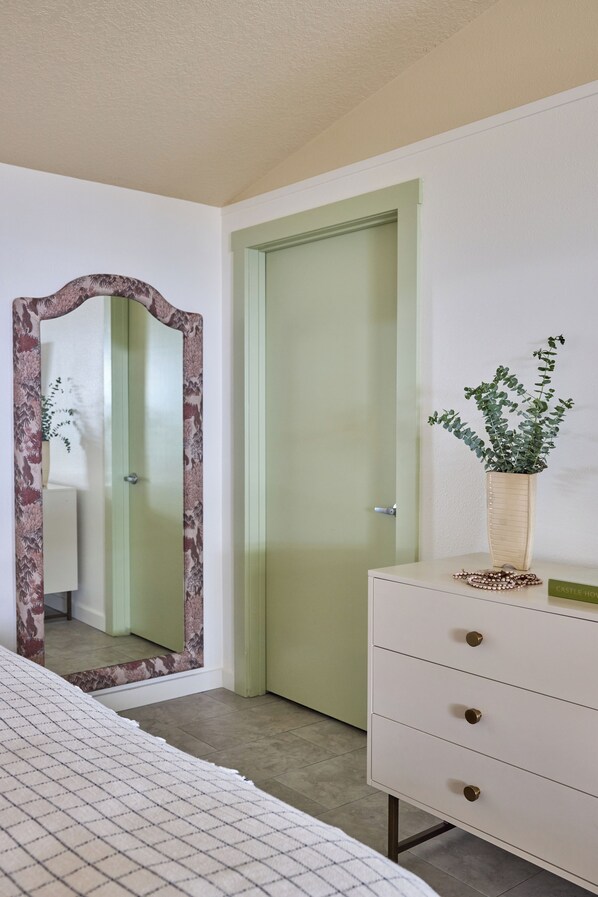 A bedroom with a bed, a dresser, a mirror with a floral frame, a green door, and a potted plant.