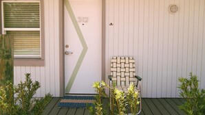 A door with the number 312, a window with blinds, a green door mat, a chair with a geometric pattern, and small plants. - Hotel Palms (Atlantic Beach)