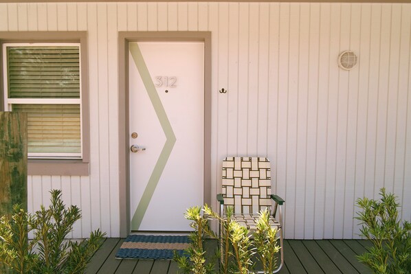A door with the number 312, a window with blinds, a green door mat, a chair with a geometric pattern, and small plants. - Hotel Palms (Atlantic Beach)
