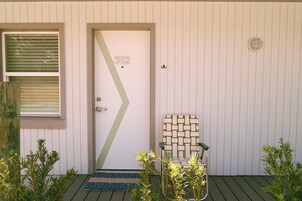 Una puerta con el número 312, una ventana con persianas, un felpudo verde, una silla con estampado geométrico y pequeñas plantas.