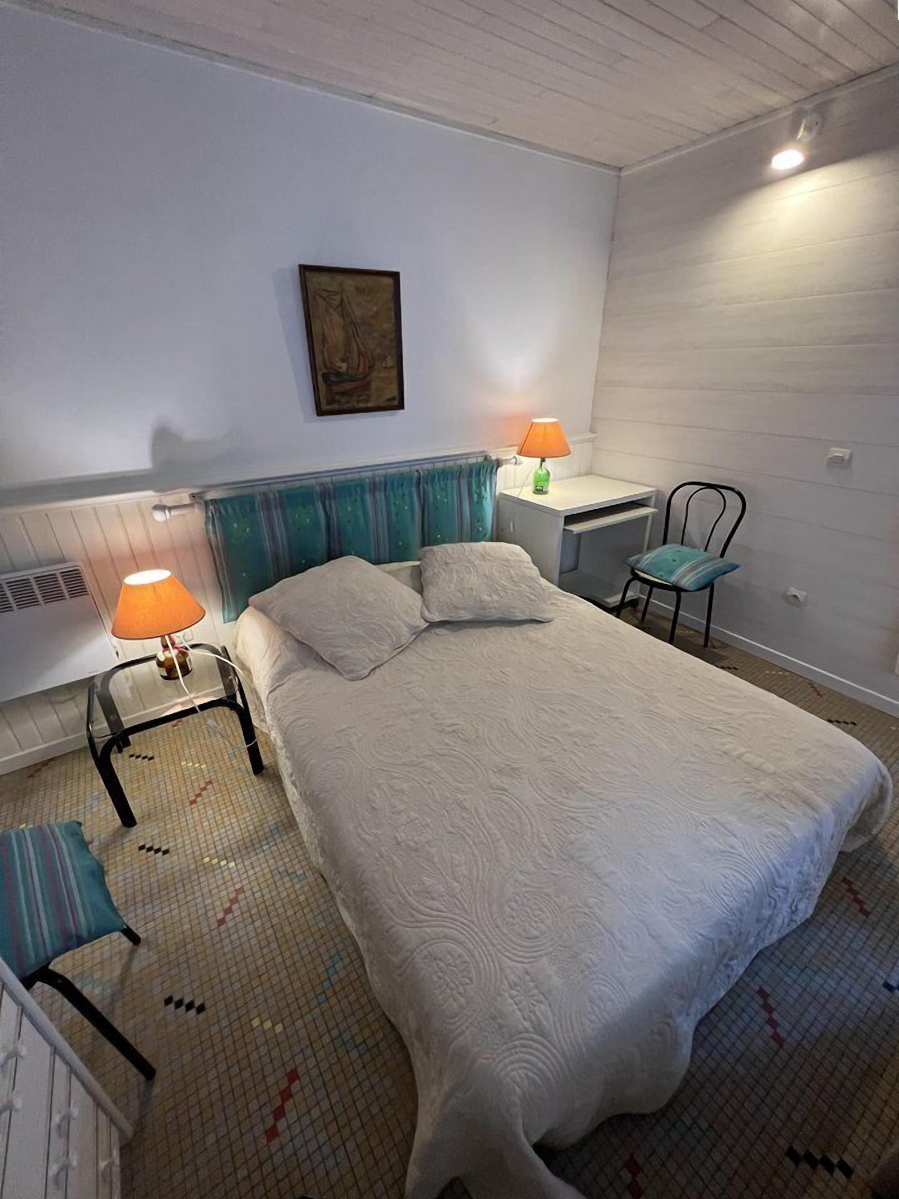 Ein ordentlich eingerichtetes Schlafzimmer mit einem Bett, zwei Nachttischlampen, einem Sessel und einem Schreibtisch.