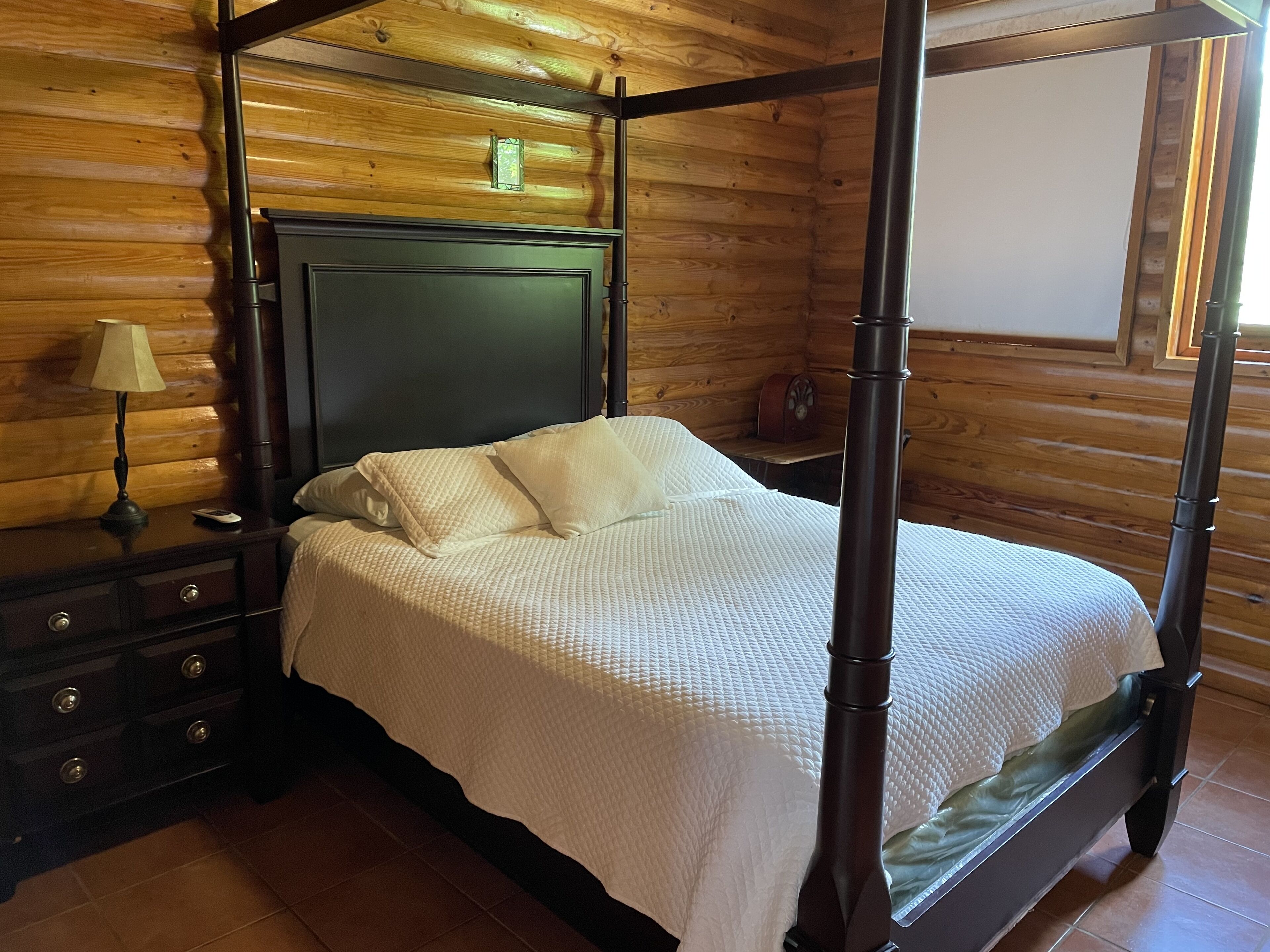 A four-poster bed with a dark headboard, a bedside table with a lamp, and a wooden wall with a clock.