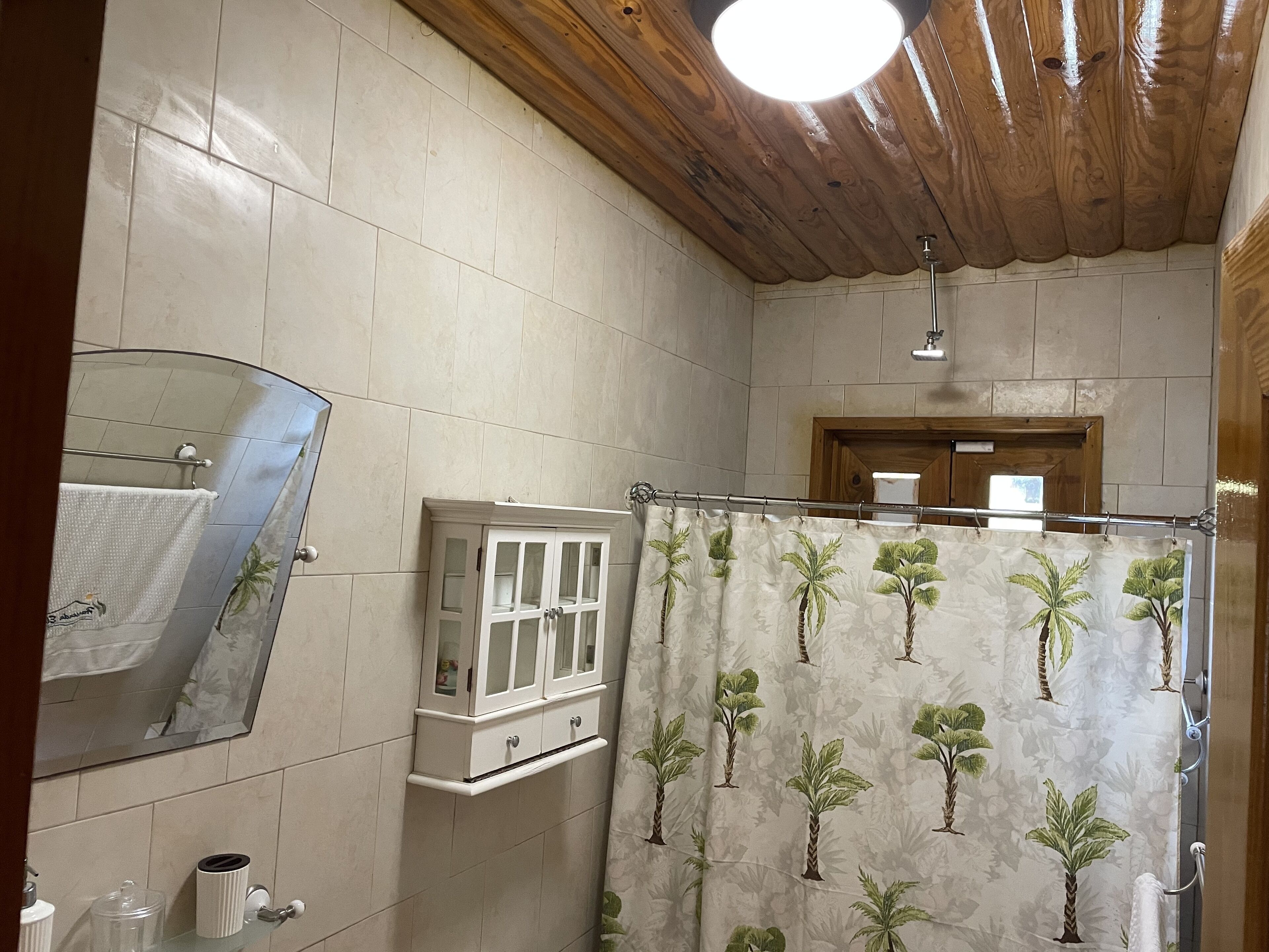 Un baño con cortina de ducha estampada con palmeras, techo de madera y espejo con una toalla colgando.