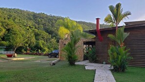 A wooden cabin with a stone foundation, a red chimney, and a pathway leading to the entrance. - Hacienda Elena Paraiso Tropical (Canóvanas)