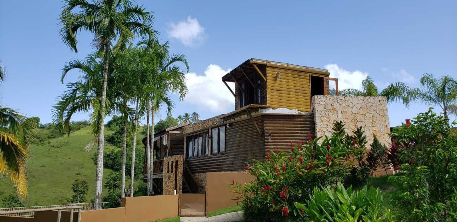 A two-story wooden house with a stone wall and a balcony, surrounded by lush greenery and palm trees.