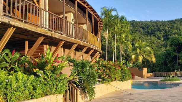 A wooden building with a balcony overlooking a pool area, surrounded by lush greenery and a forested hill in the background. - Hacienda Elena Paraiso Tropical (Canóvanas)