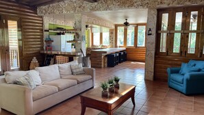 A spacious living area with a sofa, a coffee table, and a blue armchair. A kitchen area is visible in the background with a dining table and chairs. - Hacienda Elena Paraiso Tropical (Canóvanas)