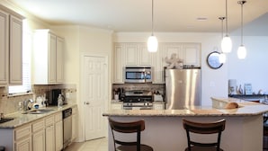 A kitchen with white cabinets, a stainless steel refrigerator, a microwave, an oven, and a sink. There are two wooden chairs at the island. - Welcome to Casa Seabrook, your By-The-Sea-Get-Away!! (Seabrook)