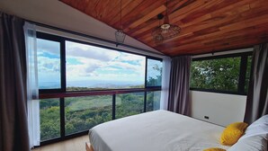 Une chambre à coucher avec un grand lit, un plafond en bois et une vue sur une forêt, visible à travers de grandes fenêtres.