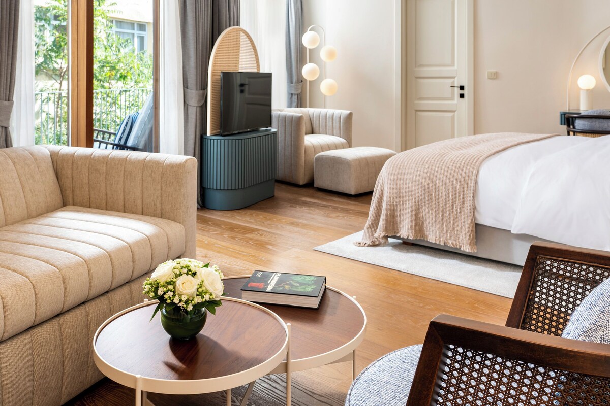 A hotel room with a sofa, a bed, a TV, a small table with a vase of flowers, and a chair.