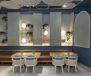 Een modern interieur met een zithoek, bestaande uit ronde tafels en rieten stoelen, een wand met verticale panelen en hangplanten, en een plafond met opvallende verlichtingsarmaturen.