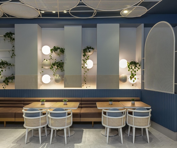 Un intérieur moderne avec un espace salon comprenant des tables rondes et des chaises en osier, un mur avec des panneaux verticaux et des plantes suspendues, et un plafond agrémenté de luminaires originaux.