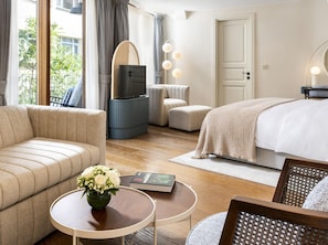 Une chambre d’hôtel avec un canapé, un lit, une télévision, une petite table avec un livre et des fleurs, et une chaise.