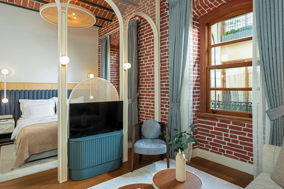 Une chambre moderne avec un mur en briques, une grande fenêtre, un lit, un téléviseur et un coin salon comprenant une chaise et une petite table.
