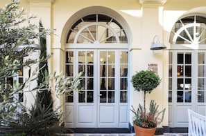 Une entrée d’immeuble avec des portes blanches et des fenêtres en arc de cercle, un pot de fleurs et une enseigne indiquant un restaurant.