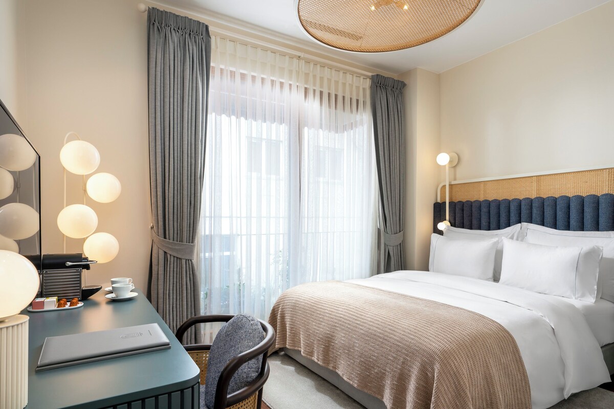 A hotel room with a bed, a desk with a laptop, a chair, and a large window with curtains.