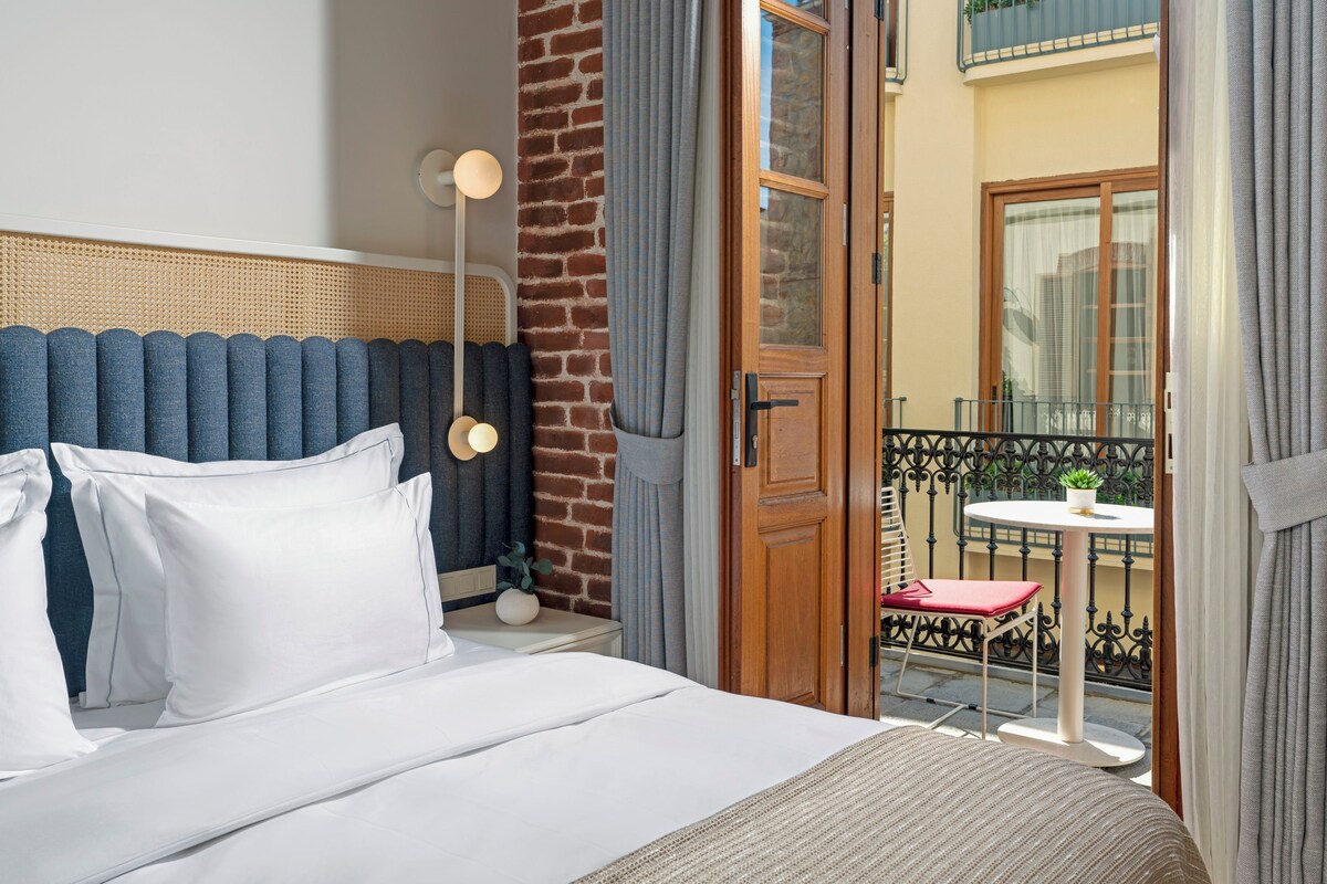 A hotel room with a bed, a nightstand, a lamp, a window with curtains, and a view of the outside balcony.