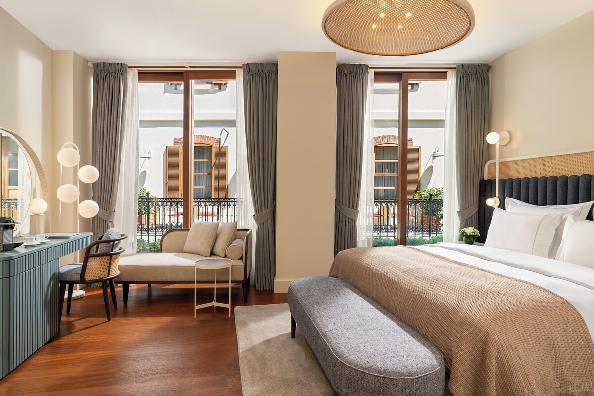 A modern hotel room with a large bed, a desk with a chair, a sofa, and a balcony with a view of buildings.