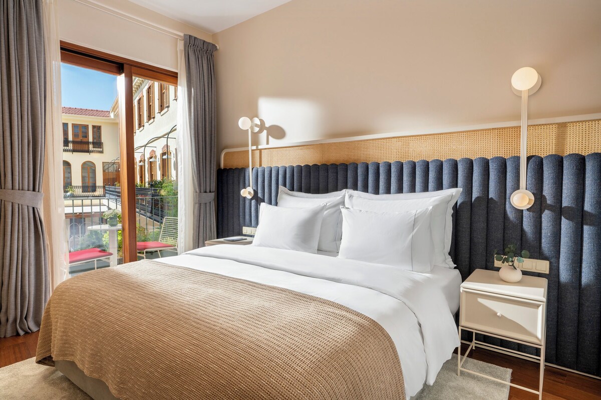 A hotel room with a large bed, a bedside table, and a view of a balcony with a railing and plants.