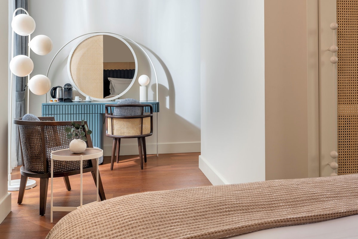 A bedroom with a bed, a vanity with a mirror, two wicker chairs, a small table with a kettle, and a blue dresser.