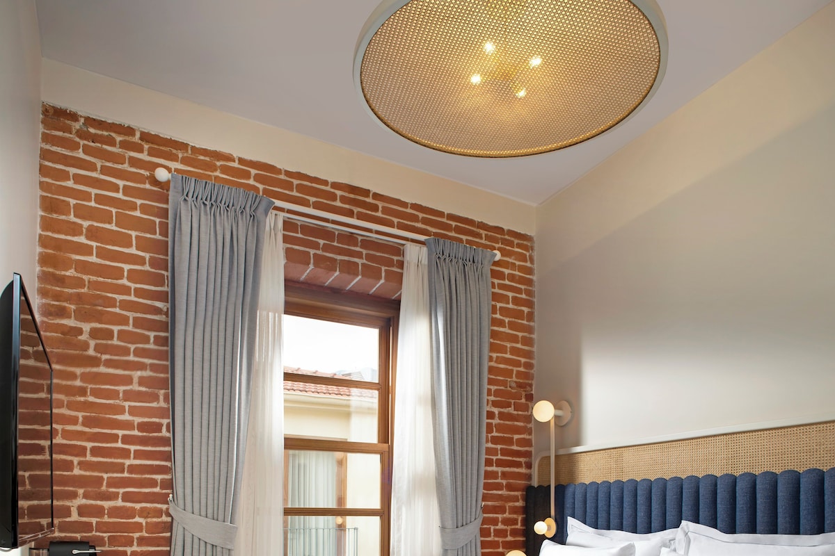 A bedroom with a brick wall, a large window with curtains, a bed with white linens, and a round ceiling light.