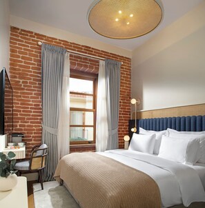 Une chambre d’hôtel avec un mur en briques, une grande fenêtre avec des rideaux, un lit avec du linge de lit blanc, une table de chevet avec une lampe et un téléviseur.