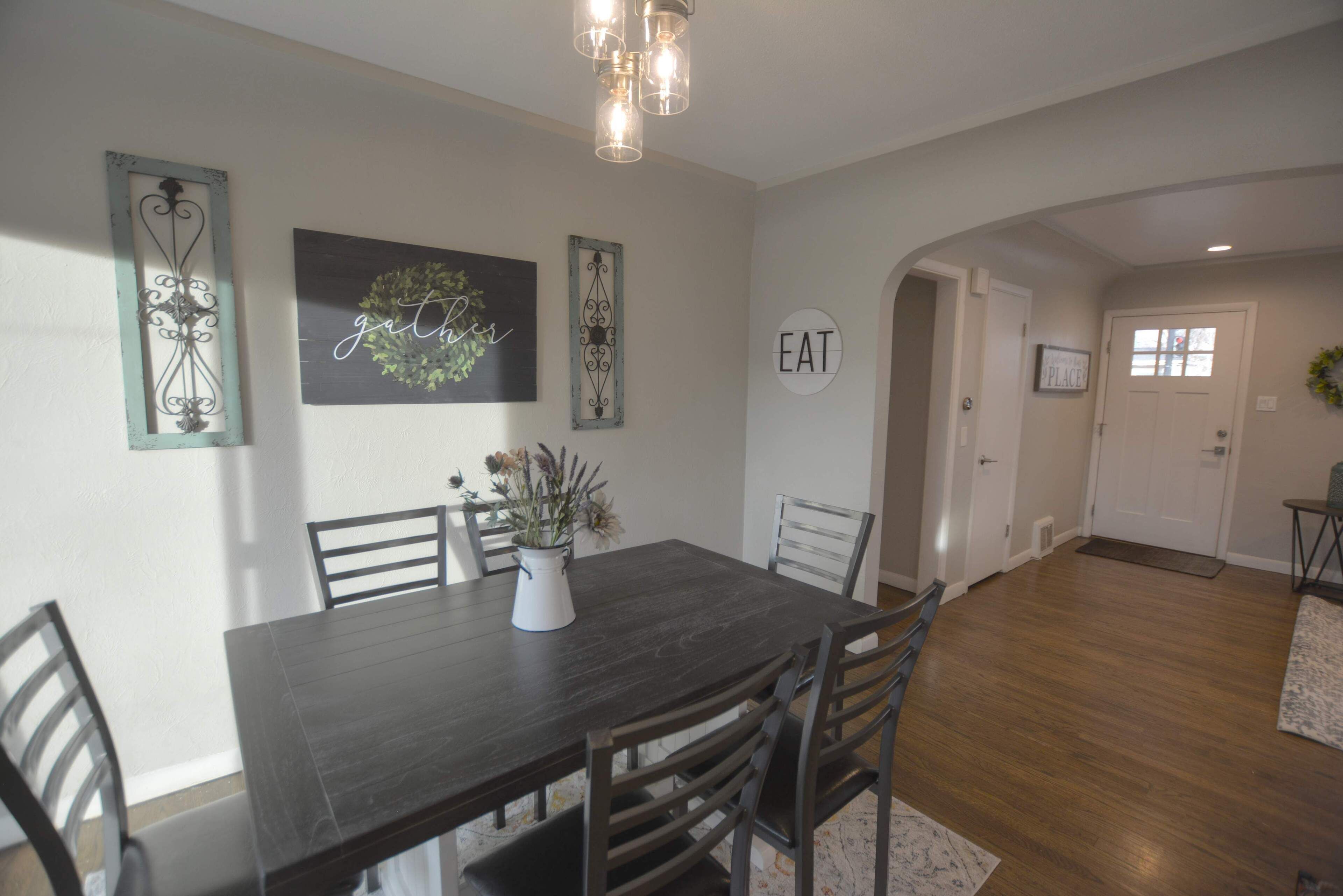 A dining area with a dark wooden table, six chairs, a vase with flowers, and wall decorations including a 'gather' sign and a 'EAT' sign.