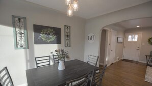 Een eetgedeelte met een donker houten tafel, zes stoelen, een vaas met bloemen en wanddecoraties, waaronder een bord met de tekst 'gather' en een bord met de tekst 'EAT'.