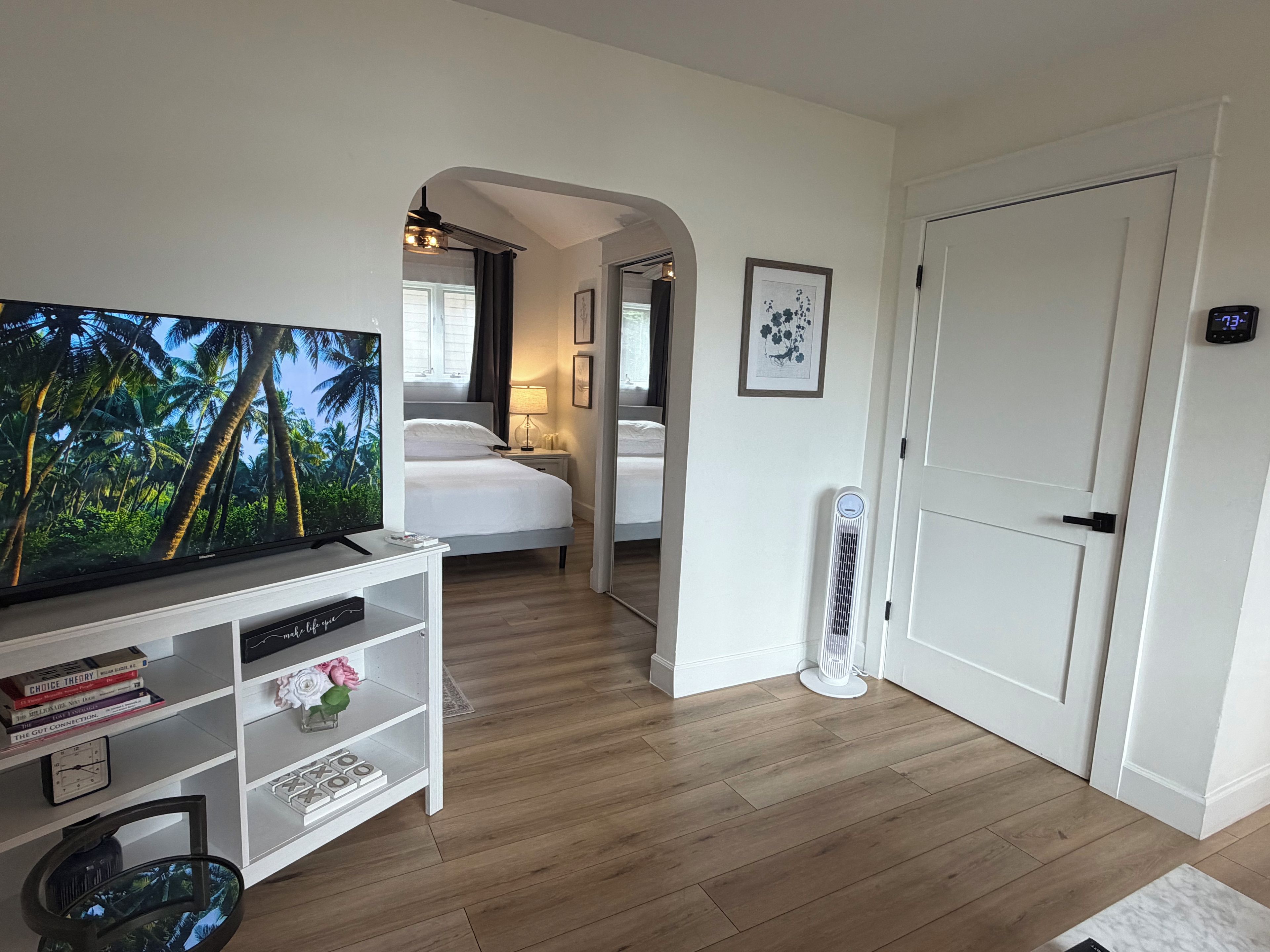 Een hotelkamer met een televisie, een boekenkast, een bed, een nachtkastje, een spiegel, een deur en een wandthermostaat.