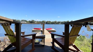 Una terraza de madera con dos mecedoras y una mesa, con vistas a un lago tranquilo con un muelle y cojines rojos.