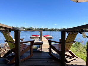 Un pontile di legno con due poltrone a dondolo e un tavolo, affacciato su un lago calmo con un molo e cuscini rossi.