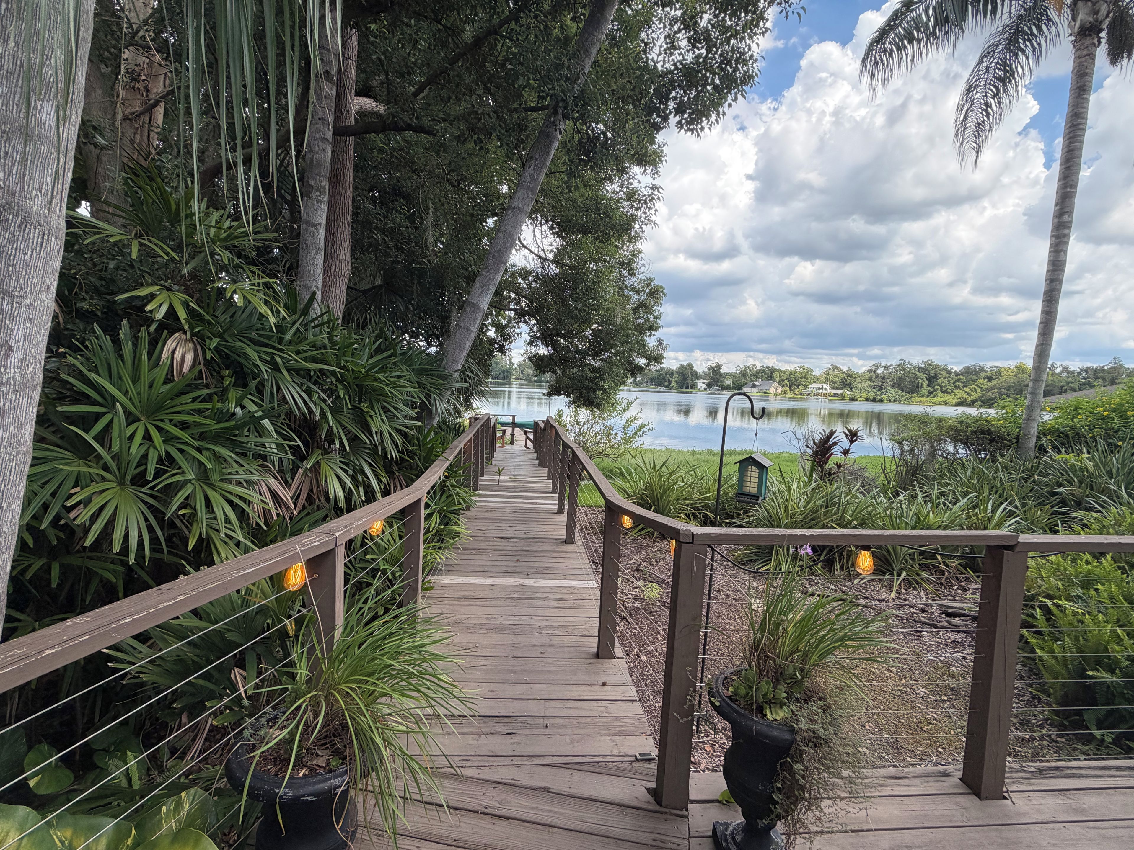 Un paseo marítimo de madera que lleva a un lago con un pabellón y plantas en macetas.