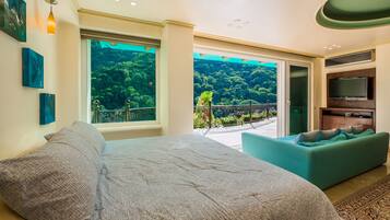 A bedroom with a large bed, a sofa, a TV, and a balcony with a view of a forest.