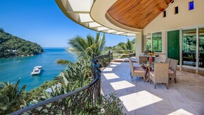 A balcony with a view of a coastal area, a dining area with wicker furniture, and a wooden ceiling. - P V South Shore Luxury Accommodations (Puerto Vallarta)