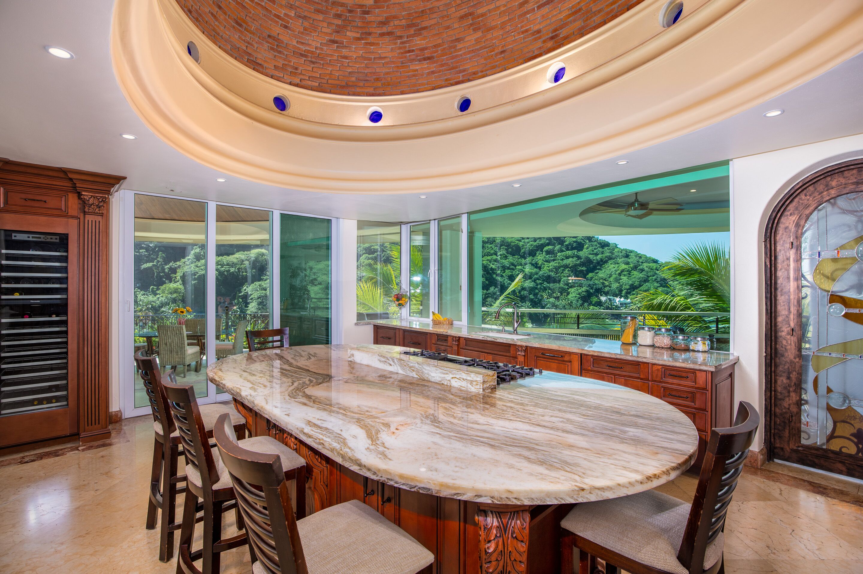 Una cocina amplia con una isla grande de mármol, armarios de madera y ventanas grandes que ofrecen vistas de un follaje exuberante.