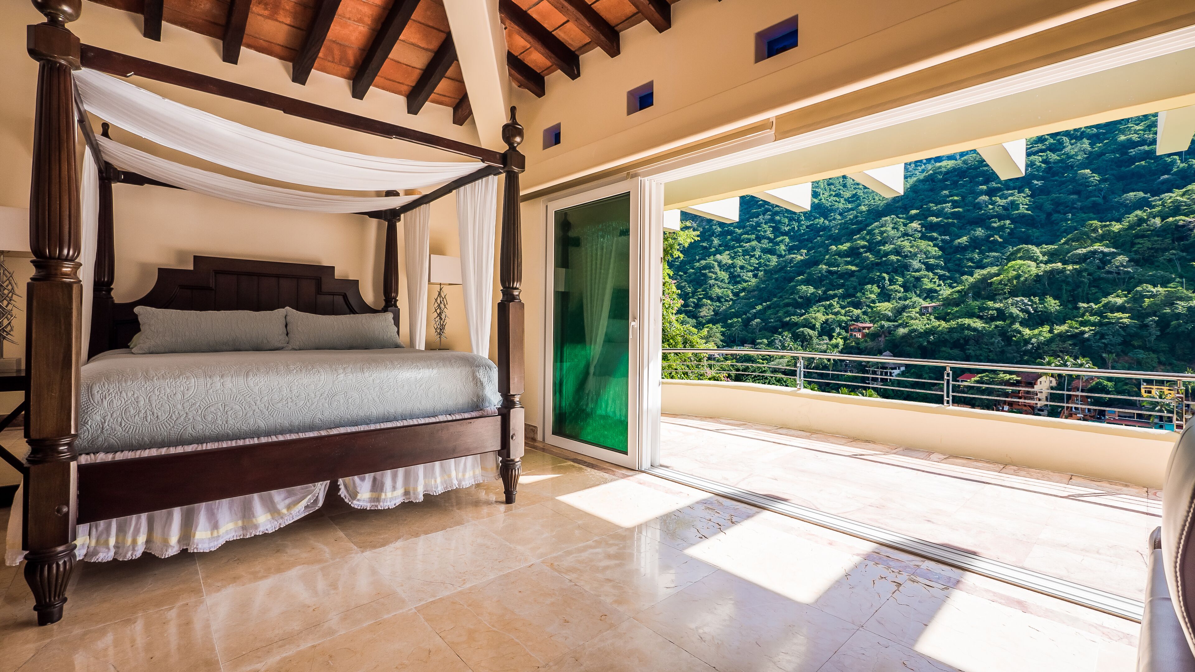 Ein Bett mit Baldachin, ein großes Fenster mit Blick auf einen bewaldeten Hang und ein Balkon mit Geländer.