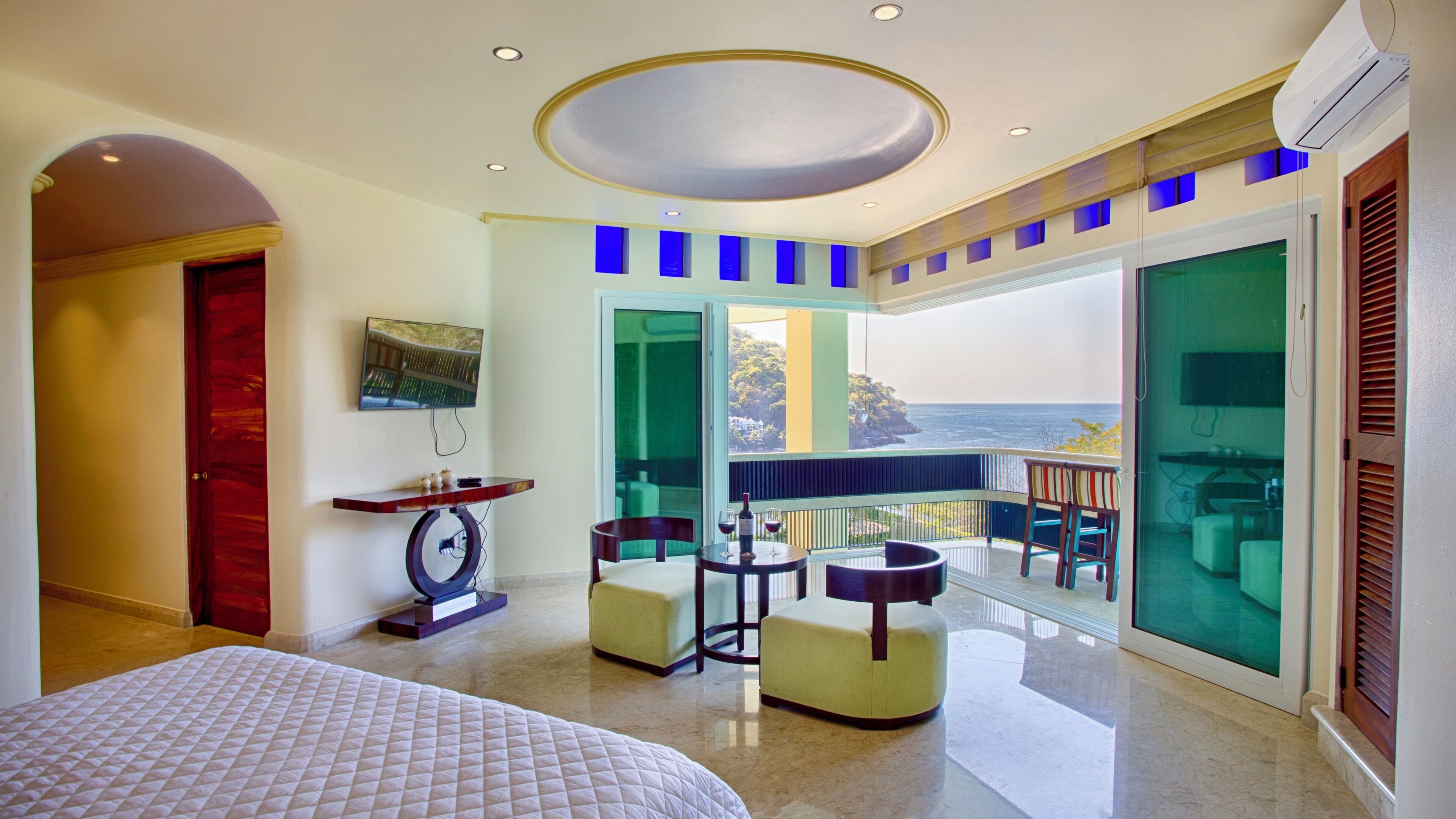 A hotel room with a bed, a small table with a vase and wine glasses, a TV on the wall, and a balcony with a view of the ocean.