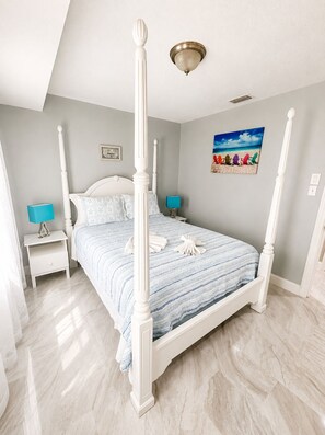 A four-poster bed with a striped bedspread, two bedside tables with lamps, a ceiling light fixture, and a beach-themed painting on the wall.