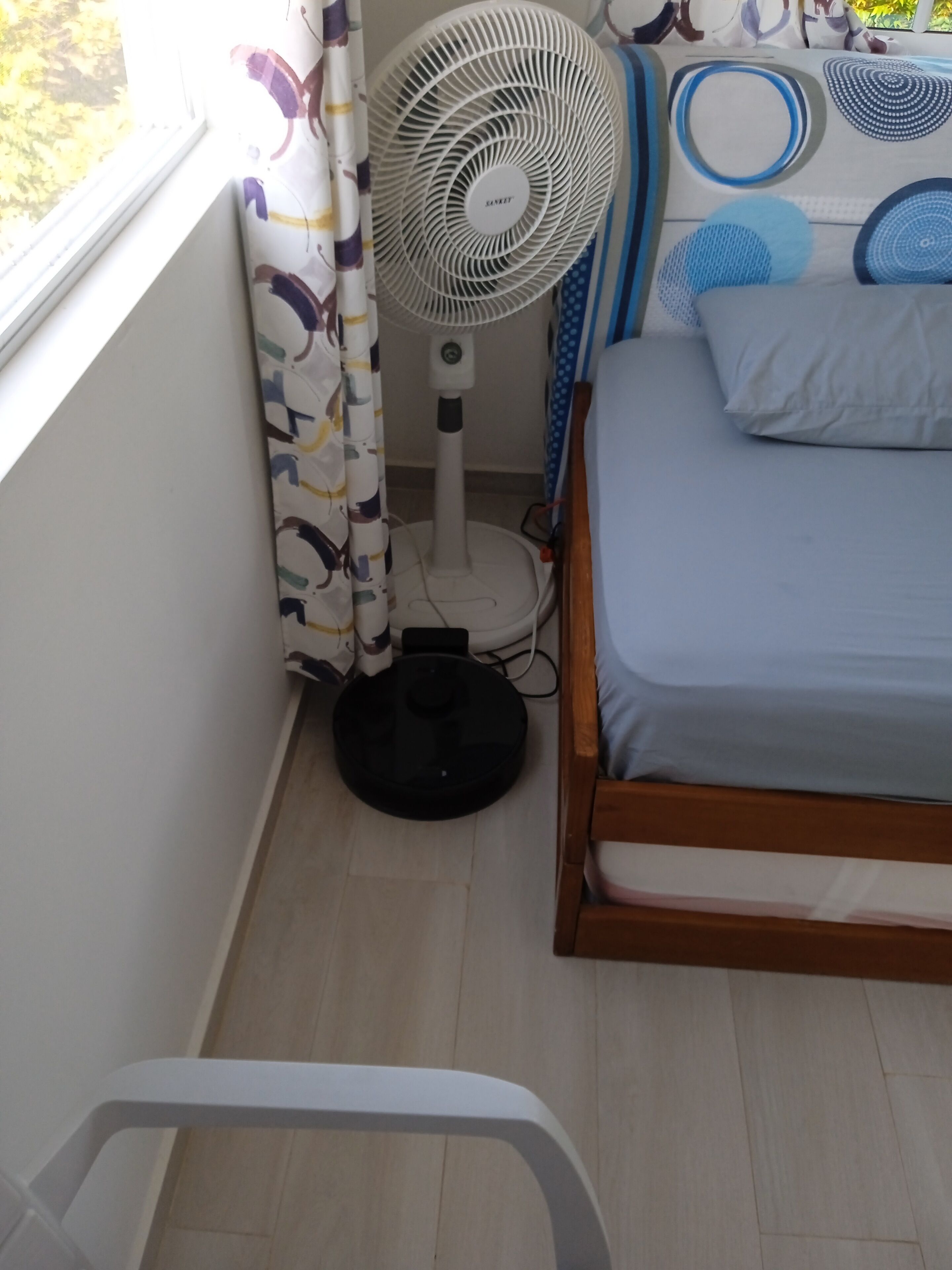 Ein kleines Zimmer mit einem Bett, einem Standventilator und einem Fenster mit Vorhängen.