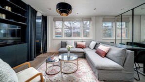 Ein modernes Wohnzimmer mit einem grauen Ecksofa, einem runden Couchtisch, einer schwarzen Hängeleuchte und einem großen Fenster mit vielen kleinen Scheiben.