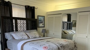 A bedroom with a dark wooden bed, a white duvet with Paris-themed prints, a small bedside table with a lamp, and a large mirror on the wall.