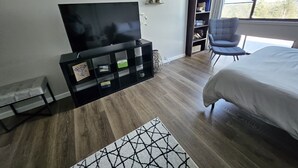 A modern bedroom with a large bed, a TV on a black stand, a bookshelf, a chair, and a window with a view of trees.