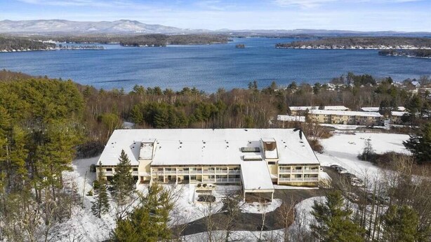 A large building with multiple sections, surrounded by snow-covered ground and trees, overlooking a large body of water.