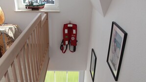 Un escalier avec des rampes en bois mène à un palier. Un petit modèle de navire est posé sur un rebord de fenêtre, et un extincteur est fixé au mur.